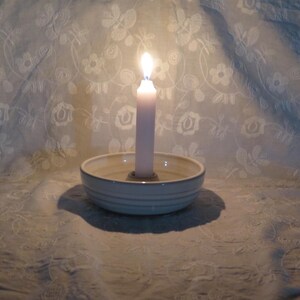 May include: A white ceramic candle holder with a circular base and a central socket, holding a single lit white taper candle. The candle emits a warm glow, illuminating the smooth, rounded edges of the bowl-shaped holder and its interior.