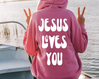 May include: A pink hooded sweatshirt with the text "JESUS LOVES YOU" in white, large, bubble letters on the back. The person wearing the sweatshirt is making a peace sign with their fingers.