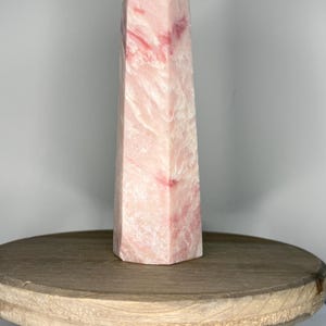 May include: A tall, pink quartz obelisk with red veining. The crystal is a light pink color with a polished surface and a pointed top. The obelisk is standing on a small, round wooden table.