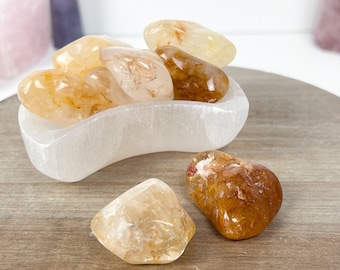 May include: A collection of tumbled citrine crystals in a white selenite bowl. The citrine crystals are a light yellow color with some orange hues. The crystals are smooth and polished.