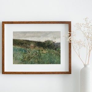 May include: A framed landscape painting with a brown wooden frame. The artwork depicts a field of green grass and wildflowers, with a path leading into the distance. The sky is overcast. A white vase with dried flowers is to the right.