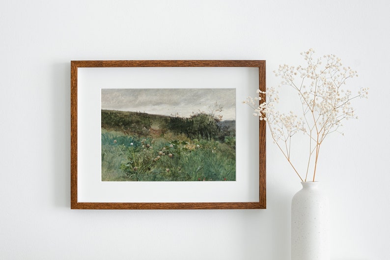 May include: A framed landscape painting with a brown wooden frame. The artwork depicts a field of green grass and wildflowers, with a path leading into the distance. The sky is overcast. A white vase with dried flowers is to the right.