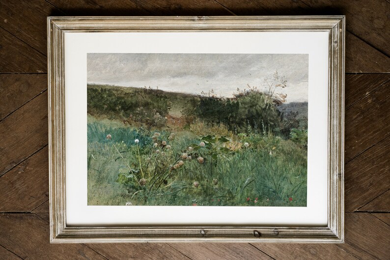 May include: A framed landscape print featuring a field of tall green grass and wildflowers under a cloudy sky. The artwork is in a silver-toned frame, adding a touch of elegance to the natural scene. The print evokes a sense of tranquility.