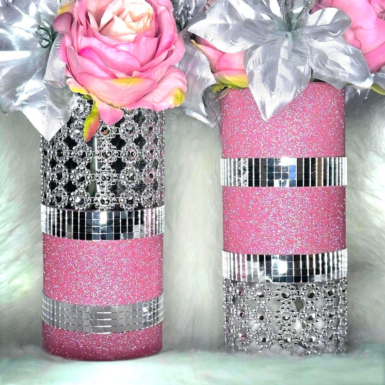 May include: Two cylindrical vases decorated with pink glitter, silver mirrored tiles, and silver floral-shaped rhinestone mesh. The vases are topped with large artificial pink roses and silver leaves, creating a decorative floral arrangement.