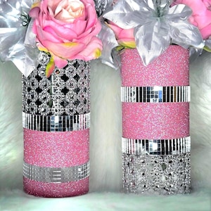 May include: Two cylindrical vases decorated with pink glitter, silver mirrored mosaic tiles, and silver floral-shaped rhinestone mesh. The vases are filled with artificial pink roses and silver leaves, creating a textured and reflective appearance.