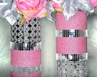 May include: Two cylindrical vases decorated with pink glitter, silver mirrored mosaic tiles, and silver floral-shaped rhinestone mesh. The vases are filled with artificial pink roses and silver leaves, creating a textured and reflective appearance.