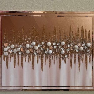 May include: A gold and white abstract art piece with a mirrored frame. The art piece features a row of white and pink beads and glitter.