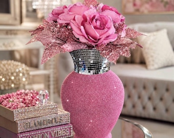 May include: A pink glitter vase with a silver mirrored neck holding a bouquet of pink roses and glittery leaves. Beside the vase are two stacked rectangular boxes with the text GLAMOROUS and FASHION VIBES, both featuring crystal-like accents.