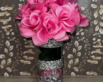 May include: A clear glass vase with silver glitter bands holds a bouquet of vibrant pink roses. The vase is filled with pink candies and sits on a wooden surface, with a floral patterned background.