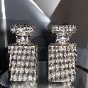 May include: Two silver perfume bottles covered in rhinestones. The bottles are square shaped and have a silver cap.