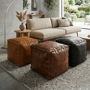 Pode incluir: Três pufe quadrados de couro em tons de castanho e preto estão dispostos sobre um tapete estampado. Um sofá bege com almofadas e uma mesa lateral de madeira com um vaso de flores secas estão ao fundo. Uma luminária de chão dourada está à esquerda.