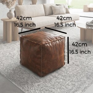 May include: A square, brown leather ottoman, 16.5 inches (42 cm) on each side, sits on a patterned rug. The ottoman is in a living room setting, with a beige sofa and wooden side tables in the background.