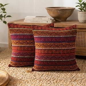 May include: Two square throw pillows featuring a horizontal striped pattern in shades of deep red, orange, purple, and gold. The fabric displays a repeating geometric motif across the bands, creating a textured appearance on the cushion covers.