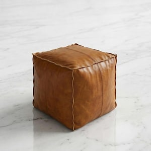 Pode incluir: Um pufe quadrado de couro castanho com costuras visíveis e uma borda bege clara. O pufe tem formato de cubo e está sobre uma superfície de mármore branco. O couro tem uma aparência desgastada.