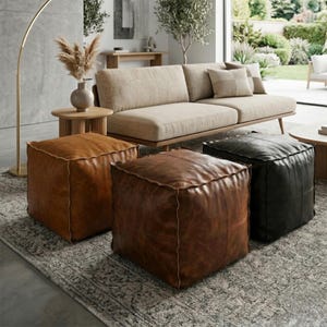 Könnte beinhalten: Drei lederne Würfelhocker in Braun- und Schwarztönen sind auf einem gemusterten Teppich angeordnet. Im Hintergrund befinden sich ein beiges Sofa mit Kissen und ein Beistelltisch aus Holz mit einer Vase mit Trockenblumen. Eine goldene Stehlampe ist ebenfalls zu sehen.