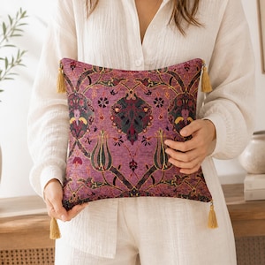 May include: A square cushion with a purple floral pattern, gold detailing, and gold tassels at each corner. The design incorporates dark green, red, and yellow elements, creating a rich, textured appearance. The cushion is a decorative item.