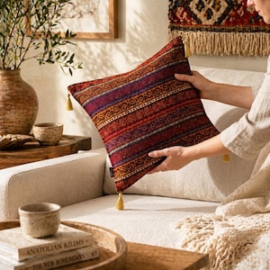 Op de afbeelding: Een decoratief vierkant kussen met een levendig, patroonontwerp in tinten rood, oranje, paars en blauw. Het kussen heeft horizontale strepen en ingewikkelde geometrische patronen. Gouden kwastjes sieren elke hoek. Het kussen ligt op een crèmekleurige bank.