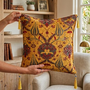 Op de afbeelding: Een decoratief vierkant kussen met een goudgele achtergrond en een gedetailleerd bloemenpatroon in grijstinten, rood en groen. Het kussen heeft decoratieve kwastjes in elke hoek, wat een vleugje elegantie toevoegt.