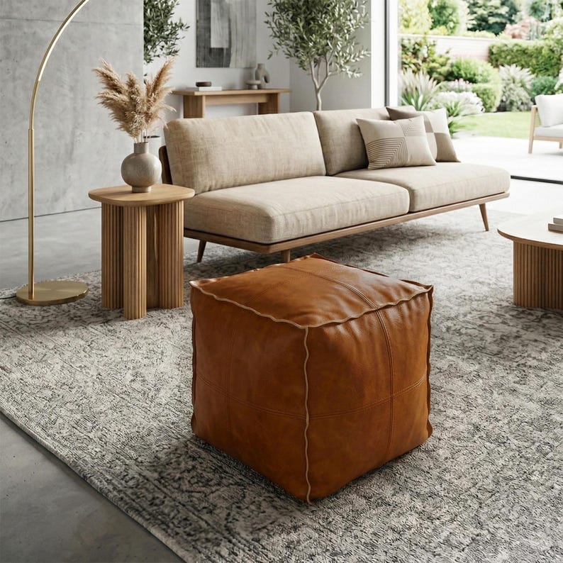 May include: A square, brown leather ottoman with light stitching sits on a patterned gray rug. The ottoman is in a living room setting with a beige sofa, wooden side tables, and a gold floor lamp. The room has a neutral color palette.