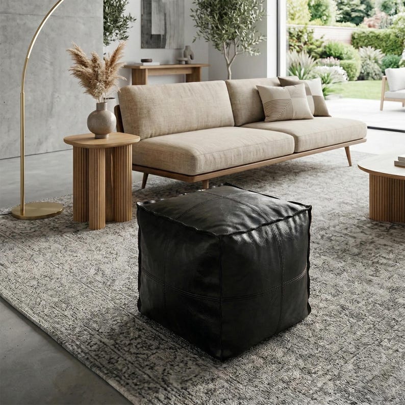 May include: A modern living room scene featuring a beige sofa with throw pillows, a black leather ottoman, and a wooden side table with a vase of dried flowers. A gold floor lamp and a patterned rug complete the decor.