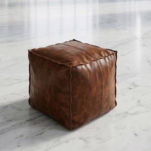 May include: A square, brown leather ottoman. The ottoman is made of stitched panels and has a distressed finish. It is a cube shape and sits on a white marble floor.