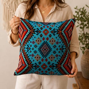 May include: A square decorative pillow with a vibrant geometric pattern. The pillow features a turquoise background with red, orange, and black tribal designs. The pillow's edges are trimmed with a zigzag pattern, adding a decorative touch. This pillow is perfect for adding a pop of color to any room.
