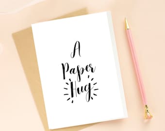 May include: A white card with black calligraphy that reads "A Paper Hug". The card is sitting on a brown envelope.