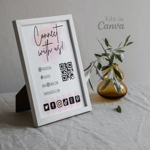 May include: A white framed sign with the text "Connect with us!" and social media icons. The sign includes a QR code and contact information. A small amber vase with greenery is in the background.