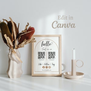 May include: A framed sign with a light brown wood frame and a white background. The sign says "hello" with a heart symbol and "Find us on" with two QR codes labeled "Facebook" and "Instagram". Below the QR codes is the text "Like | Follow | Tag" with icons for Facebook, Instagram, and Pinterest. The text "www.yoursite.com" is at the bottom of the sign.