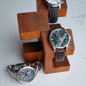 May include: Three wristwatches on a wooden watch stand. The watches have black, white, and green faces with silver bands. The stand is made of dark brown wood.