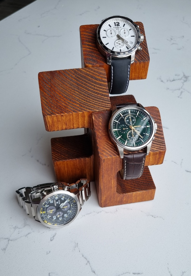 May include: Three wristwatches on a wooden watch stand. The watches have black, white, and green faces with silver bands. The stand is made of dark brown wood.
