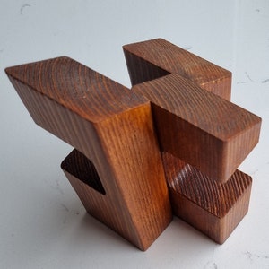 May include: A wooden puzzle made of four pieces that interlock to form a complex shape. The puzzle is made of a dark brown wood with a natural grain.