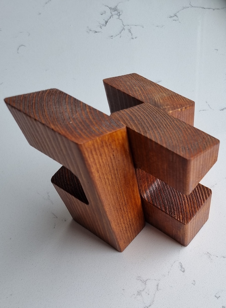 May include: A wooden puzzle made of four pieces that interlock to form a complex shape. The puzzle is made of a dark brown wood with a natural grain.