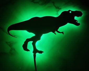 May include: A green light shines through a black silhouette of a Tyrannosaurus Rex dinosaur. The dinosaur is standing on a black metal stand.