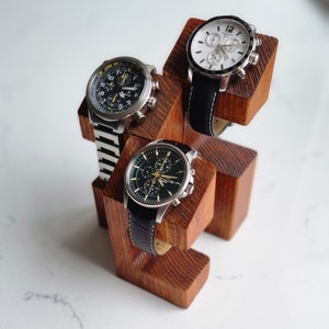 May include: A wooden watch stand with three watch slots. The stand is made of dark brown wood and has a natural finish. Three watches are displayed on the stand, two with black bands and one with a white band.