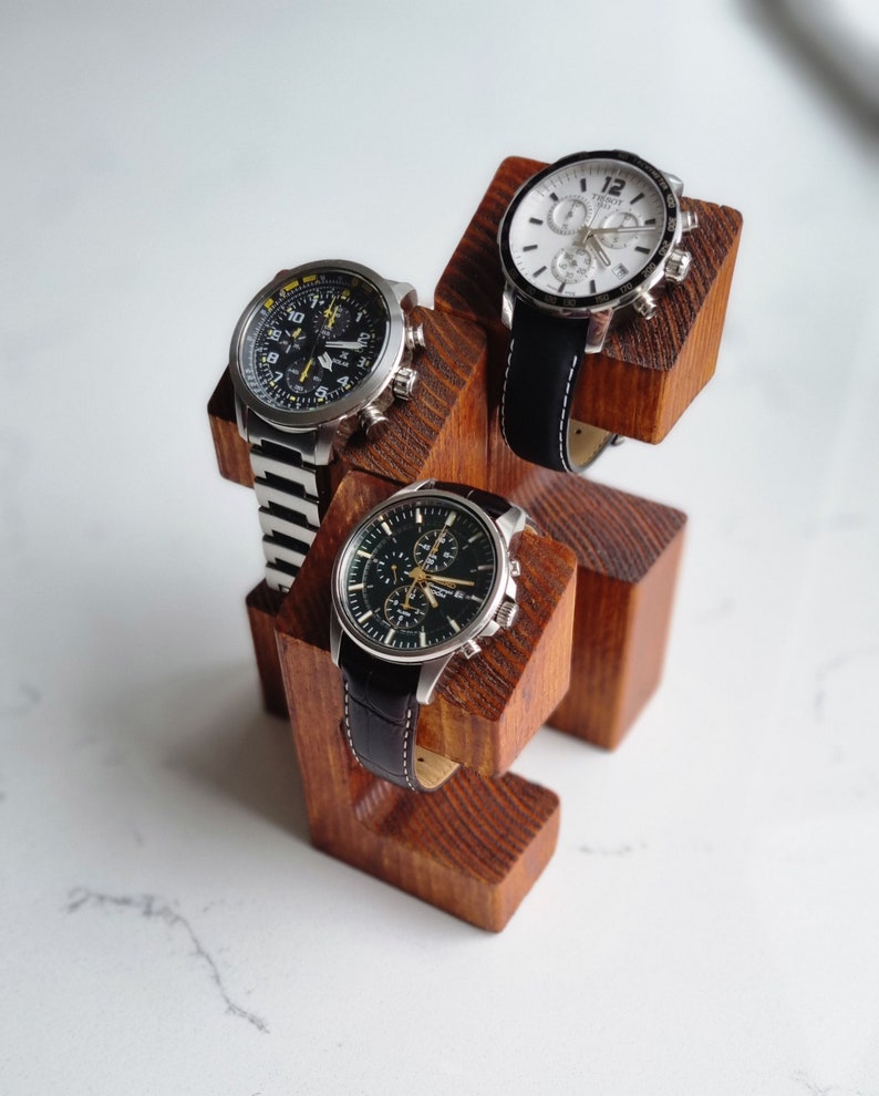 May include: A wooden watch stand with three watch slots. The stand is made of dark brown wood and has a natural finish. Three watches are displayed on the stand, two with black bands and one with a white band.