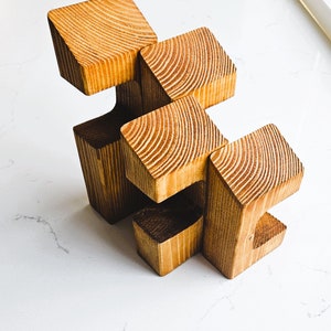 May include: A wooden sculpture made of five connected blocks. The blocks are arranged in a staggered pattern, creating a unique and abstract design. The sculpture is made of natural wood with a warm brown color.