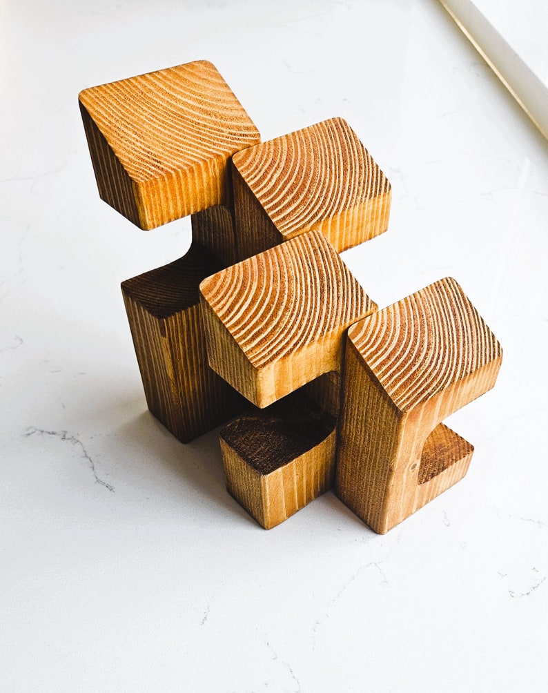 May include: A wooden sculpture made of five connected blocks. The blocks are arranged in a staggered pattern, creating a unique and abstract design. The sculpture is made of natural wood with a warm brown color.