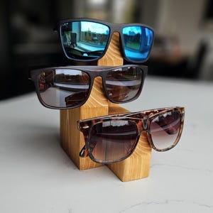 Puede incluir: Tres pares de gafas de sol expuestas en un soporte de madera. Las gafas de sol son negras con lentes espejadas azules, marrones con lentes marrones y de carey con lentes degradados marrones.
