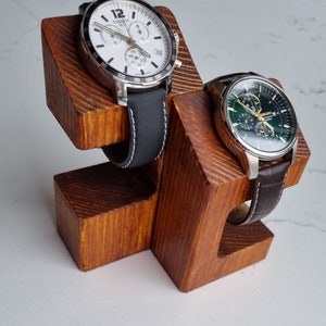 May include: A wooden watch stand with two watch slots. The stand is made of natural wood with a brown stain. The watch slots are angled to hold the watches securely. The stand is a great way to display and store your favorite watches.