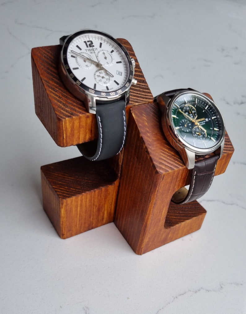 May include: A wooden watch stand with two watch slots. The stand is made of natural wood with a brown stain. The watch slots are angled to hold the watches securely. The stand is a great way to display and store your favorite watches.