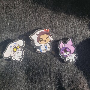Könnte beinhalten: Drei klare Acrylstifte mit Cartoon-Charakteren. Einer zeigt einen weißen, kaninchenartigen Charakter mit gelben Augen. Ein anderer zeigt einen Charakter in einem weißen Kapuzen-Outfit mit einer roten Schleife. Der dritte Stift zeigt einen lila Kapuzencharakter.