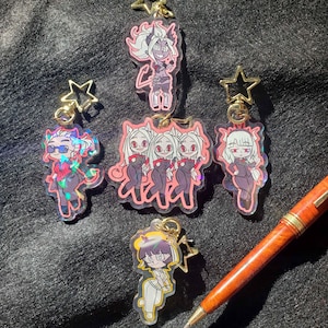 May include: A selection of acrylic keyrings with anime-style character designs. The keyrings have gold-coloured star-shaped clasps. The characters are shown in different poses and outfits, with a pink and purple colour scheme. A pen with a brown barrel is in the image.