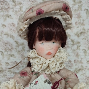 Puede incluir: Una mu&ntilde;eca artesanal con cabello casta&ntilde;o rojizo, ojos azules y pecas, vestida con un vestido con estampado floral y un gorro a juego. La mu&ntilde;eca sostiene un peque&ntilde;o libro atado con una cinta burdeos. El vestido presenta adornos de encaje y patrones de rosas.