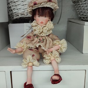Puede incluir: Una mu&ntilde;eca artesanal con est&eacute;tica vintage. La mu&ntilde;eca viste un conjunto beige con detalles florales, un sombrero a juego y sandalias rojas. El conjunto de la mu&ntilde;eca presenta adornos de encaje. La mu&ntilde;eca est&aacute; sentada sobre una superficie blanca.