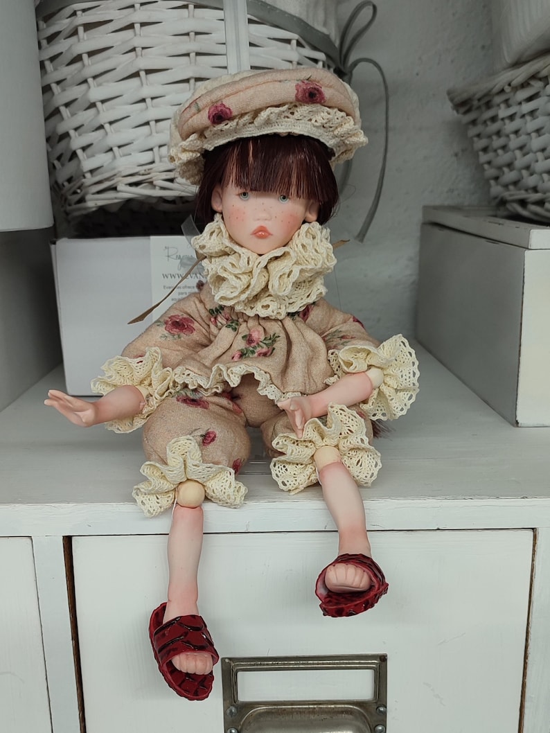 Puede incluir: Una mu&ntilde;eca artesanal con est&eacute;tica vintage. La mu&ntilde;eca viste un conjunto beige con detalles florales, un sombrero a juego y sandalias rojas. El conjunto de la mu&ntilde;eca presenta adornos de encaje. La mu&ntilde;eca est&aacute; sentada sobre una superficie blanca.