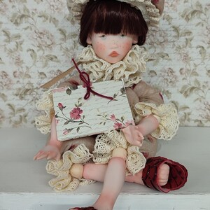 Puede incluir: Mu&ntilde;eca artesanal con expresi&oacute;n serena, vestida con un vestido beige con ribete de encaje y un gorro a juego adornado con rosas. La mu&ntilde;eca lleva sandalias rojas y sostiene una tarjeta con motivos florales.