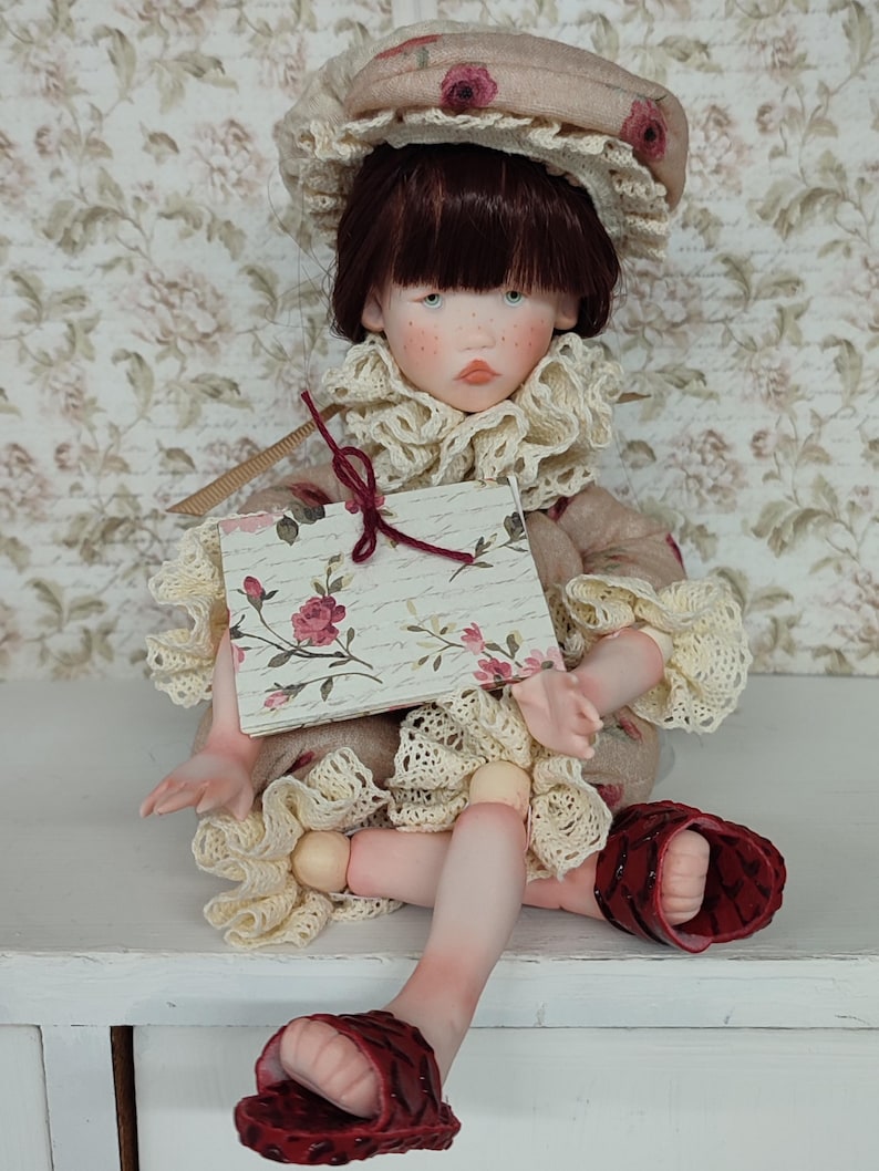 Puede incluir: Mu&ntilde;eca artesanal con expresi&oacute;n serena, vestida con un vestido beige con ribete de encaje y un gorro a juego adornado con rosas. La mu&ntilde;eca lleva sandalias rojas y sostiene una tarjeta con motivos florales.