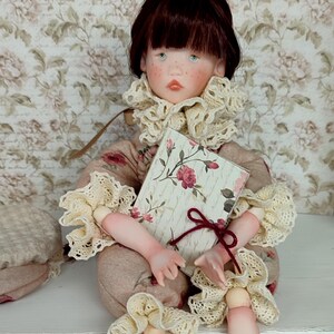 Puede incluir: Una mu&ntilde;eca artesanal con una expresi&oacute;n serena, vestida con un atuendo beige con detalles florales y adornos de encaje color crema. La mu&ntilde;eca sostiene un libro con una cubierta floral y una cinta burdeos. La mu&ntilde;eca tiene cabello casta&ntilde;o y ojos azules.
