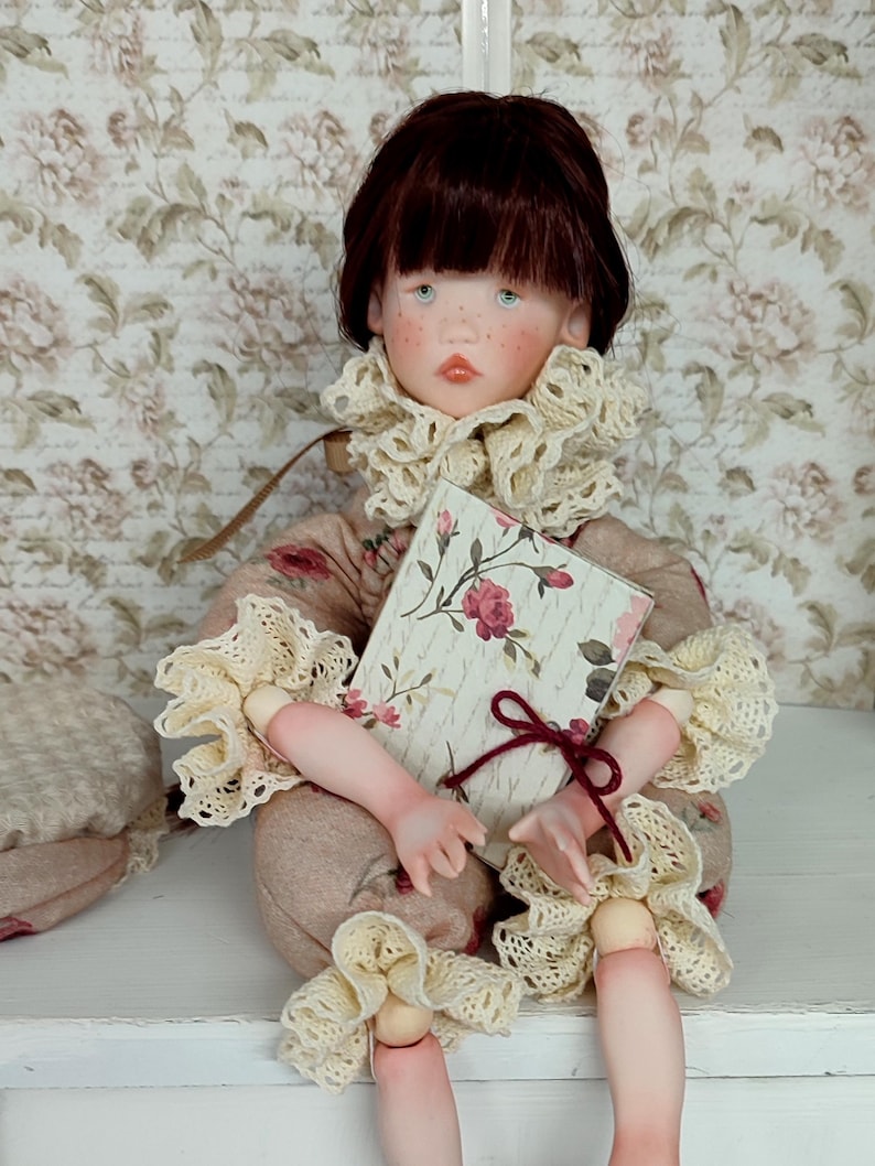 Puede incluir: Una mu&ntilde;eca artesanal con una expresi&oacute;n serena, vestida con un atuendo beige con detalles florales y adornos de encaje color crema. La mu&ntilde;eca sostiene un libro con una cubierta floral y una cinta burdeos. La mu&ntilde;eca tiene cabello casta&ntilde;o y ojos azules.